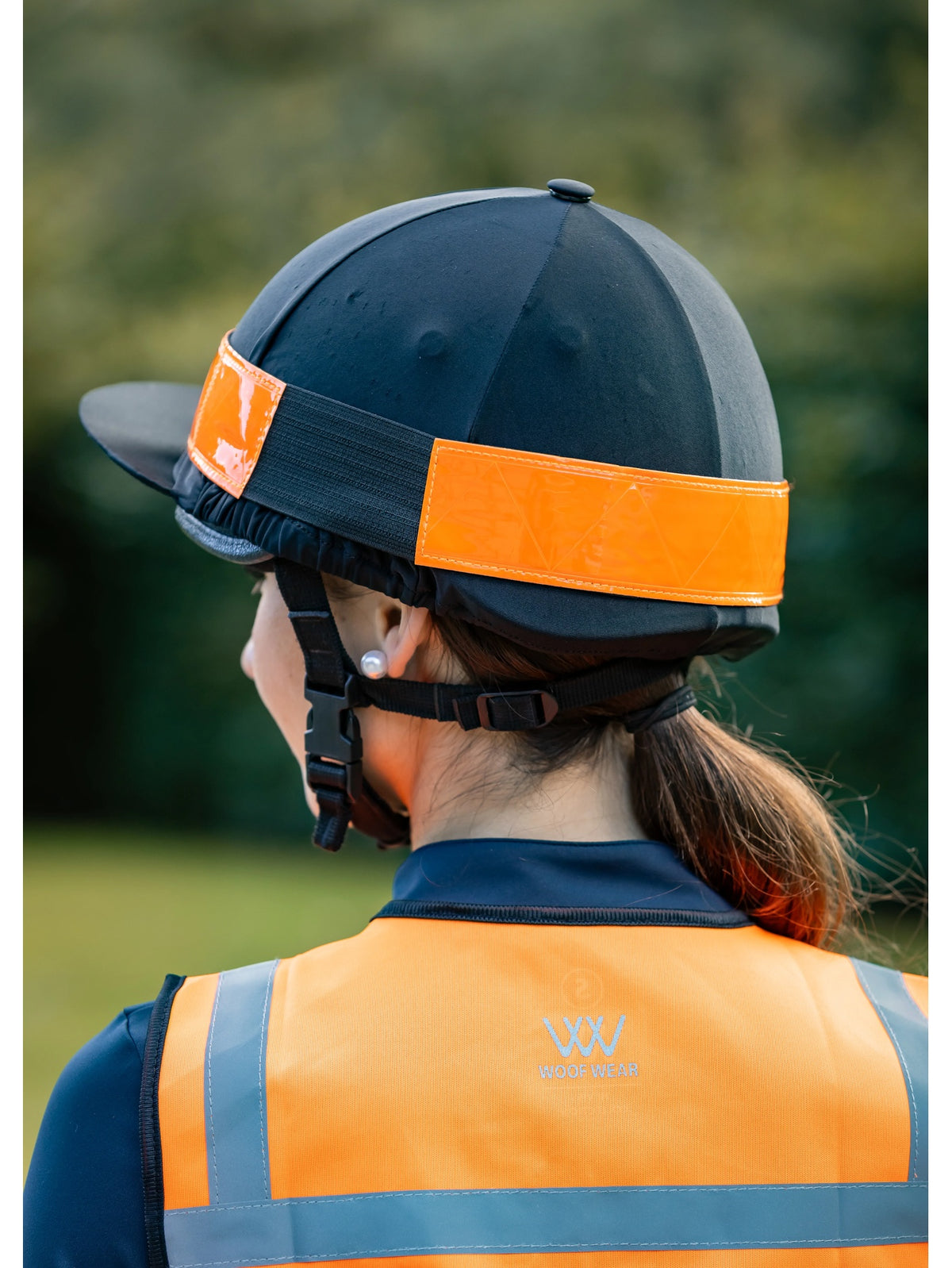 A person shown from the back wearing a black equestrian riding helmet with a bright orange reflective band and an orange high-visibility safety vest with gray reflective stripes. The background is a soft, blurred green outdoor setting.