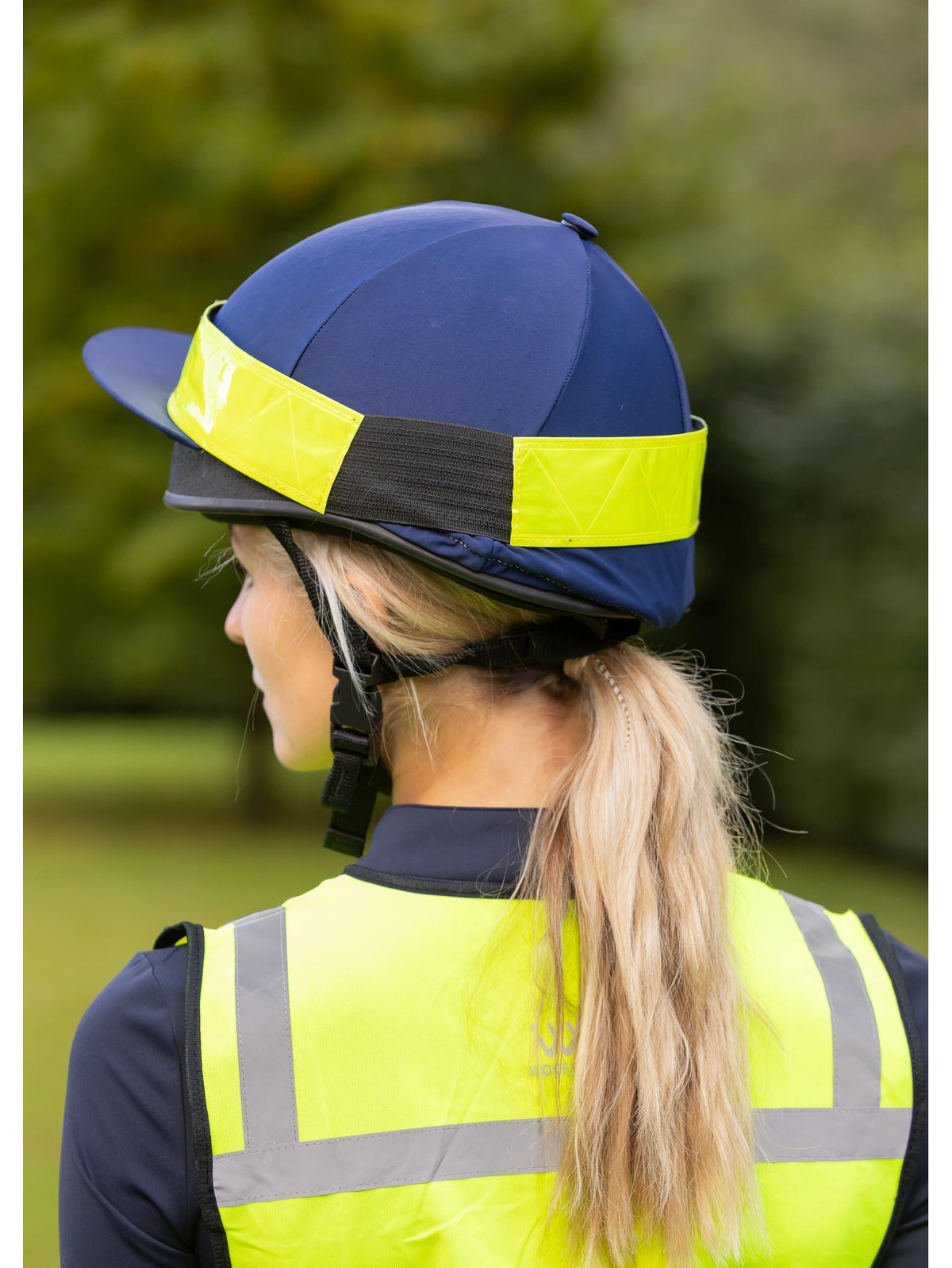 A woman wearing a navy blue riding helmet with bright yellow reflective bands and a matching high-visibility yellow safety vest, photographed outdoors with a blurred green background.