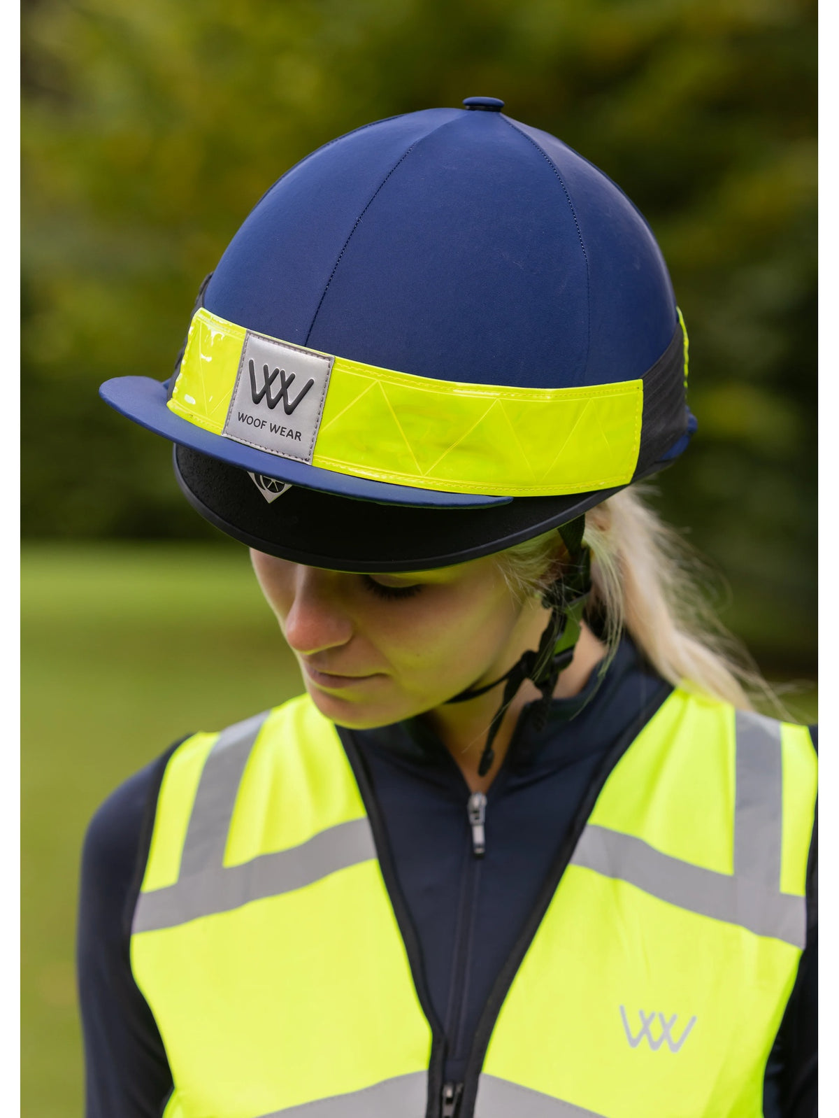 A woman outdoors wearing a navy blue riding helmet with a bright yellow reflective band and a matching high-visibility yellow vest. The helmet has a visible 'VV VVOT WEAR' logo patch on the front band.