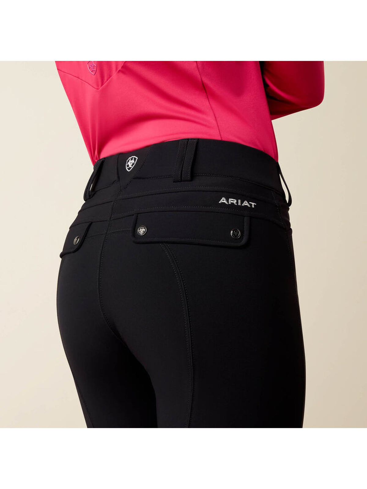Close-up rear view of a person wearing black Ariat equestrian pants with a bright pink top, against a plain light beige background.