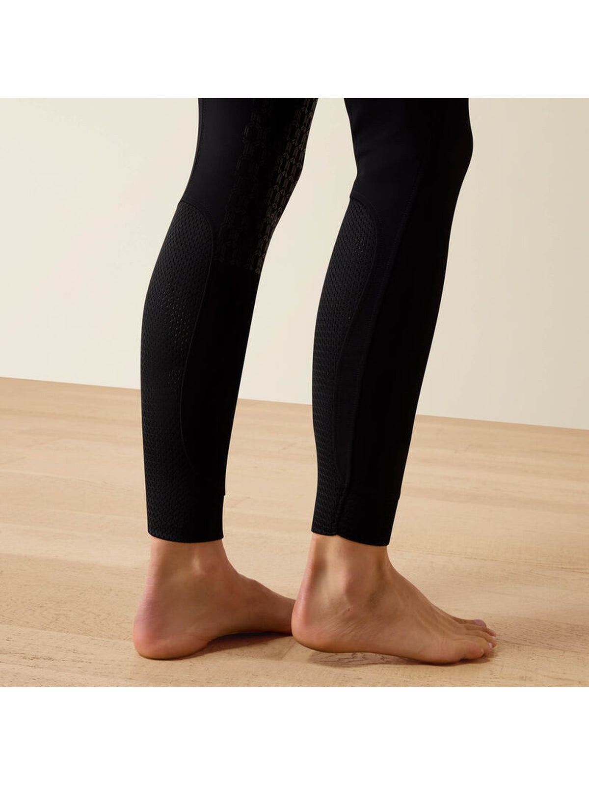Black textured athletic leggings worn by a model, shown from the back on bare feet against a light beige wall and wooden floor.